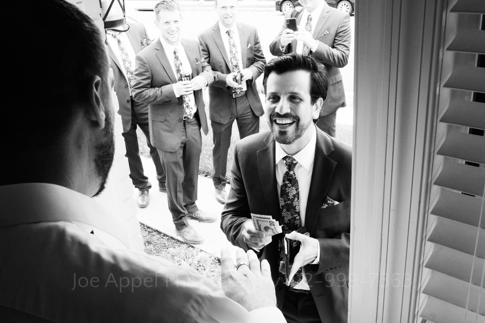 A bearded groom reaches into his wallet and pulls out money as a tall man blocking the doorway refuses the cash. Groomsmen stand behind the groom laughing.