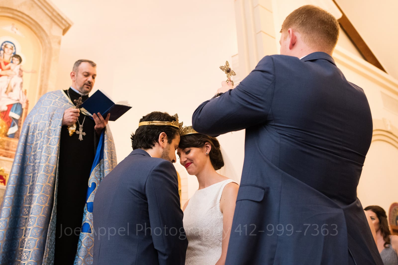 A bride and groom stand with their foreheads together while wearing crowns. A priest reads a verse from the bible above them while a man wearing a blue suit holds a crucifix while facing them at St. Mary's Armenian Apostolic Church in Washington DC.