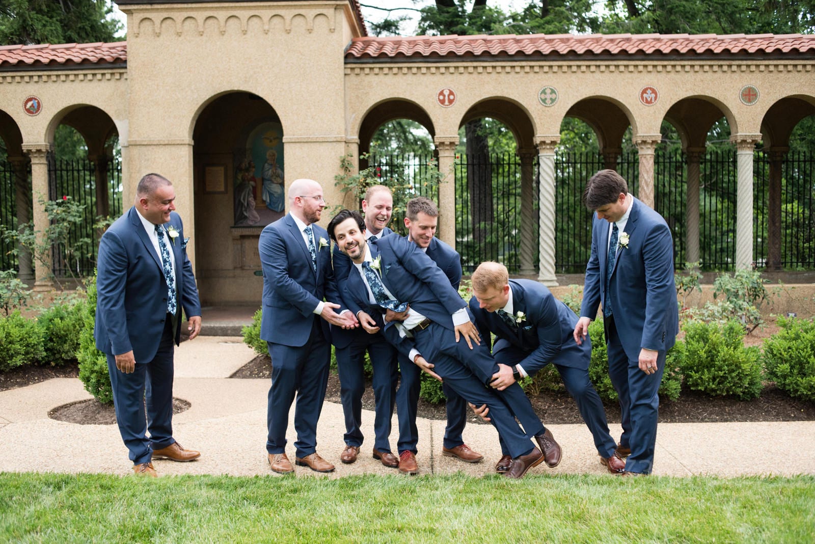 A groom holds his hands to his side as his groomsmen struggle to pick him up outside of his St Francis Hall Washington DC wedding. All the men are wearing blue suits and brown shoes.