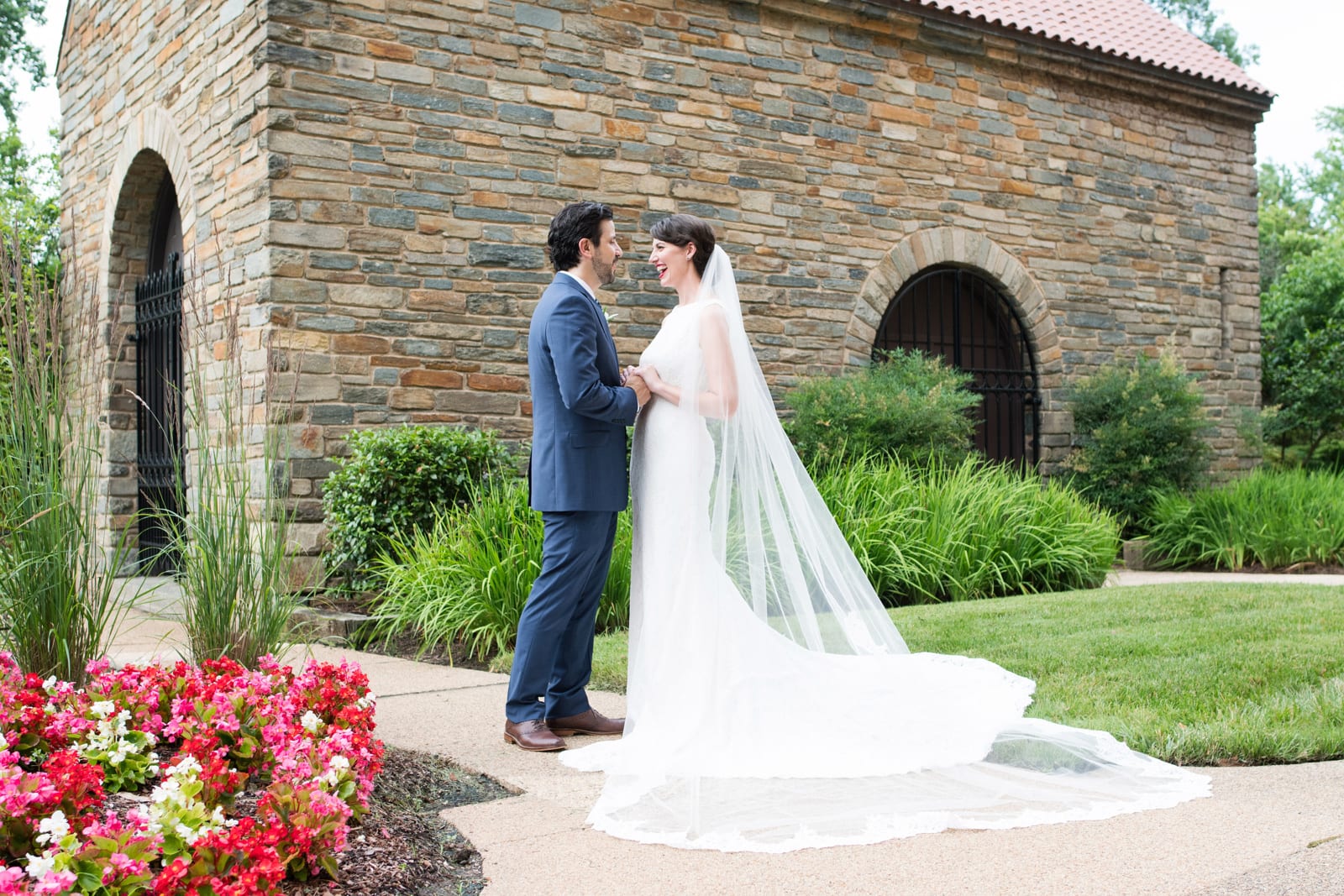 Red and pink flowers are in the foreground as a bride in a long, flowing white dress and a groom in a blue suit stand in front of a stone building with arched doorways and landscaped lawn during their St Francis Hall Washington DC wedding.