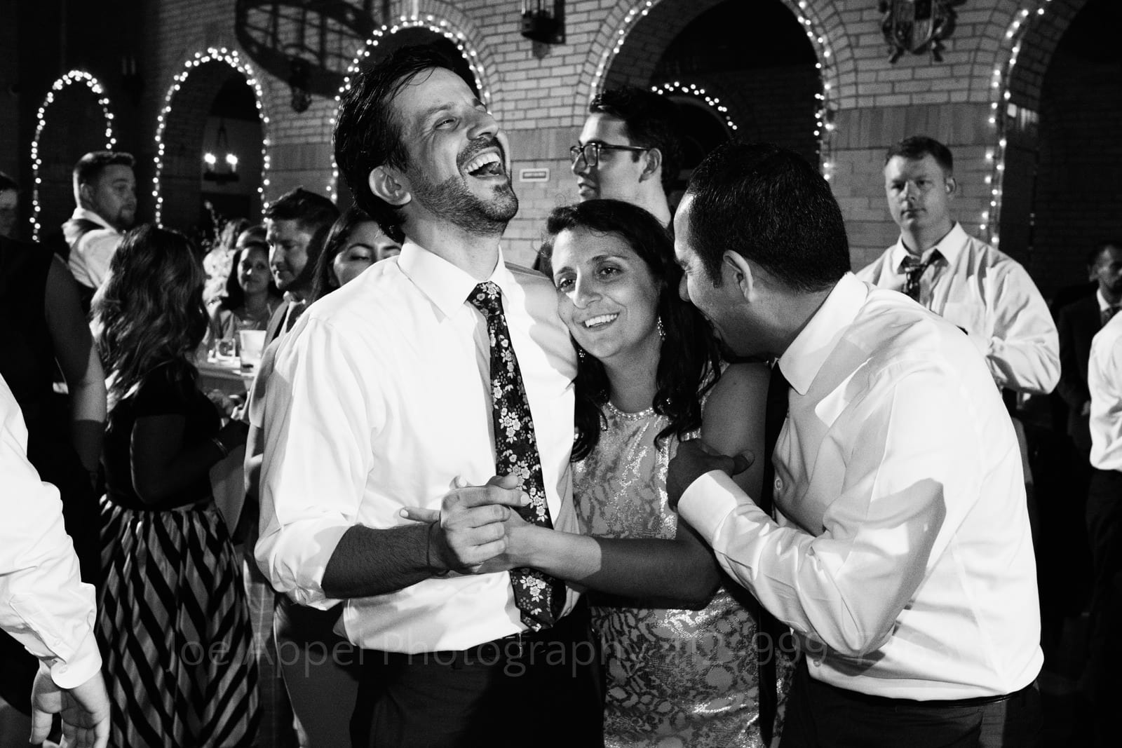 A groom in shirtsleeves laughs as he dances with his sister while another guest holds on to her and says something.