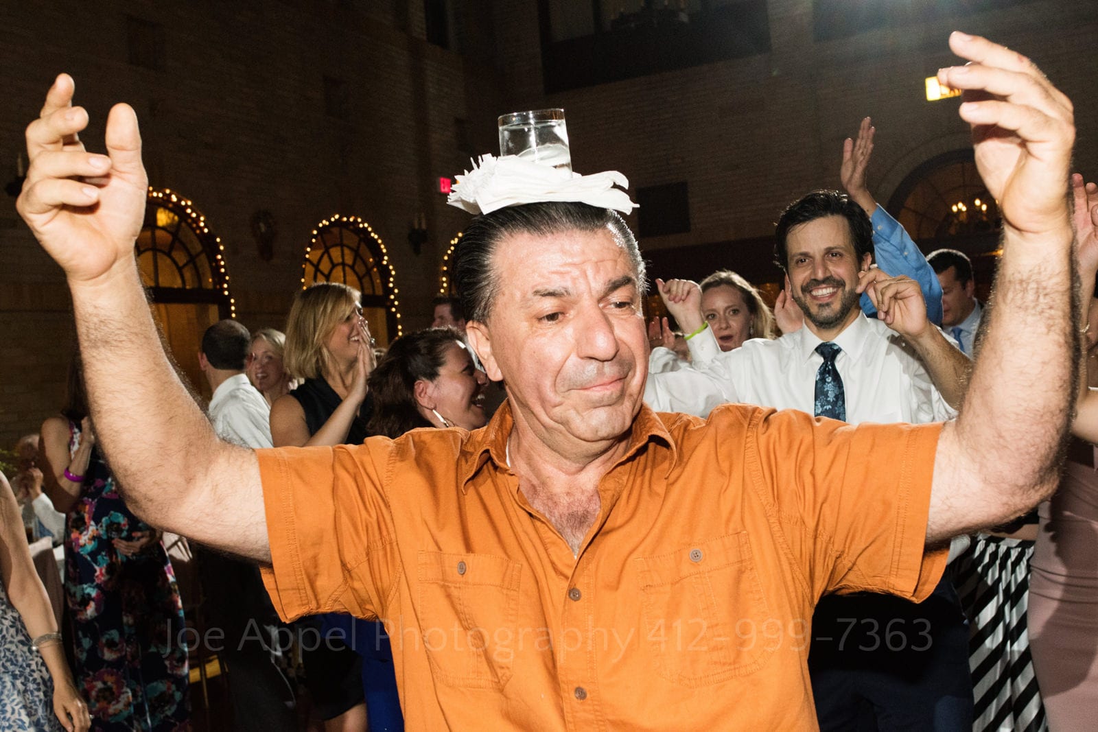 A older man in an orange shirt dances with a glass of water on his head during a St Francis Hall Washington DC wedding.