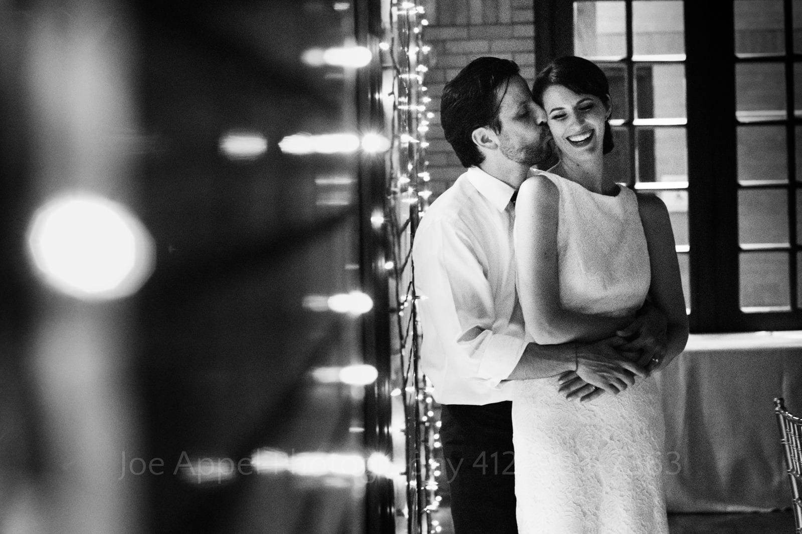 With twinkle lights in the foreground, a groom embraces his smiling bride and kisses her on the cheek during their St Francis Hall Washington DC wedding.