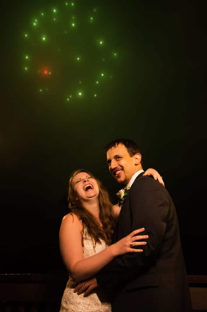 A bride and groom laugh as they embrace under a green fireworks explosion during their Fall Wedding at Seven Springs