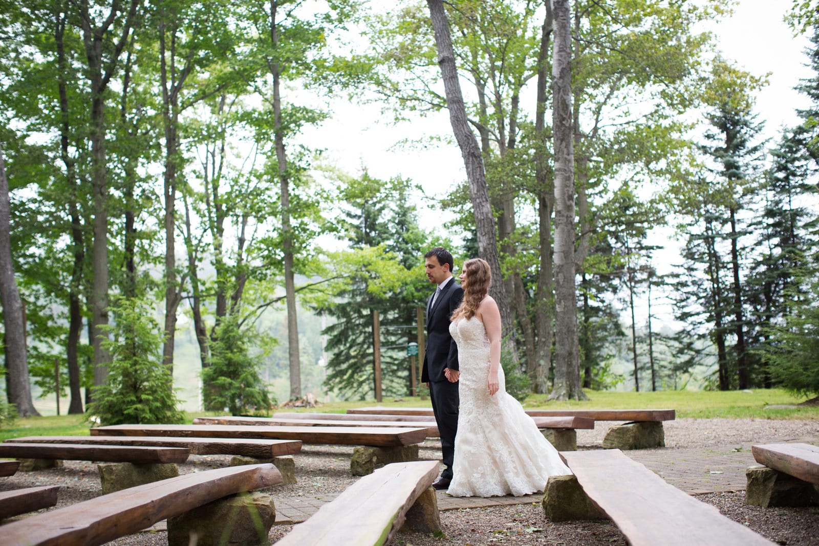 A bride and groom hold hands as they walk between two rows of benches in a wooded glade during their Fall Wedding at Seven Springs.