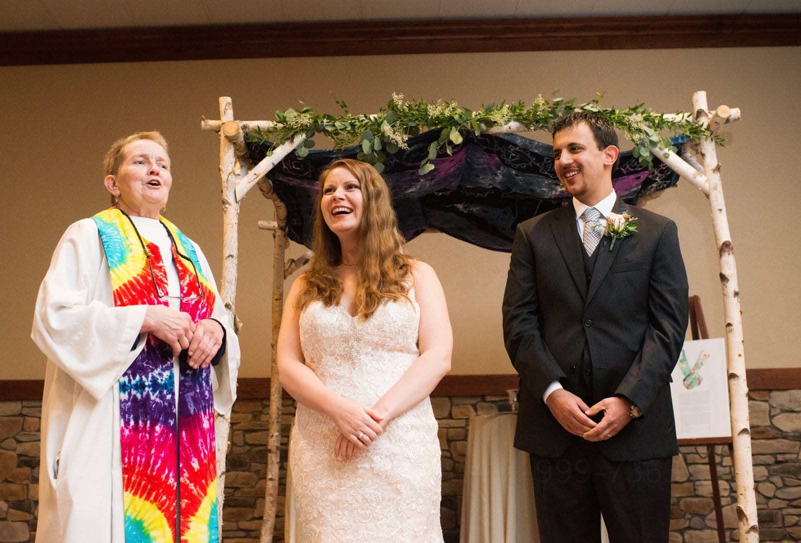 A female pastor wearing a tie dyed sash speaks as the bride and groom standing next to her smile during their Fall Wedding at Seven Springs.