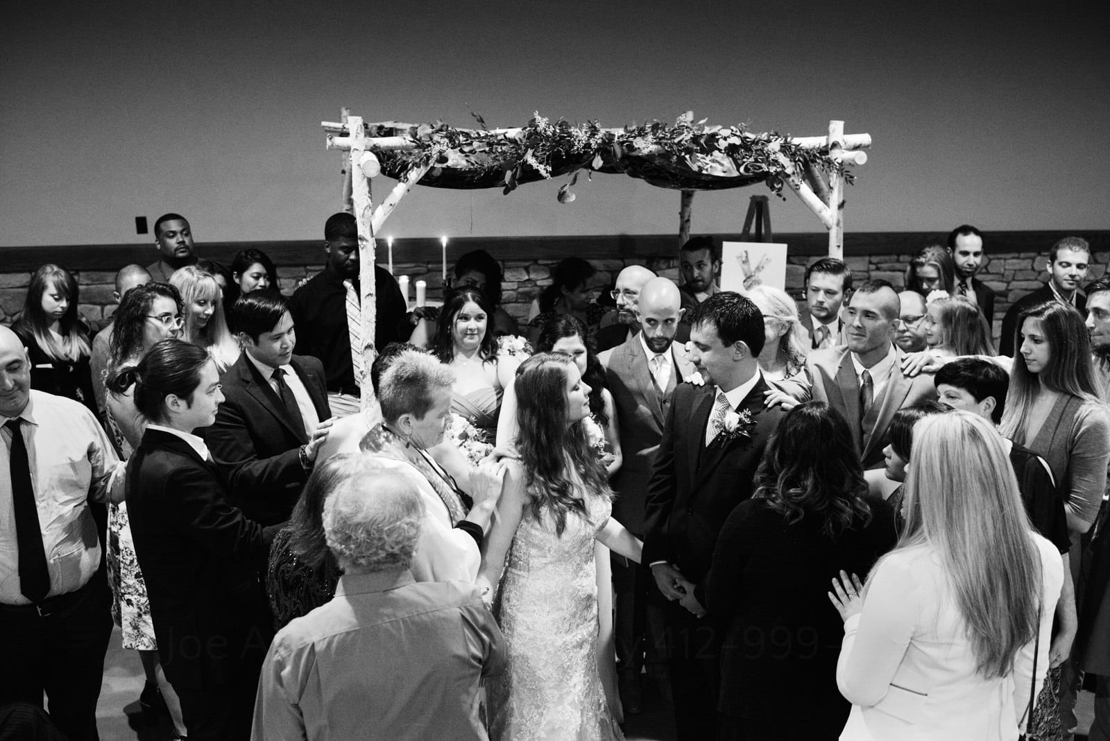 Overhead view of a bride and groom standing in the center of their wedding guests who are gathered around them everyone connected by touching each other's shoulders during their Fall Wedding at Seven Springs.
