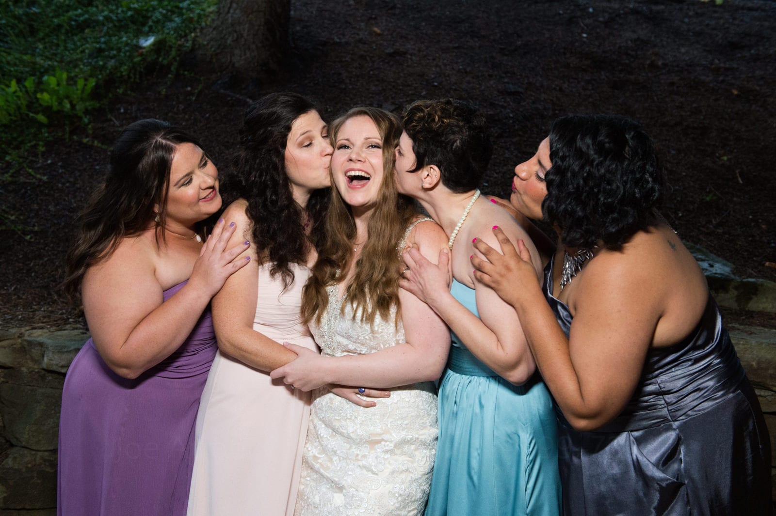A bride smiles as her four bridesmaids in blue and purple dresses hold on to her and kiss her.