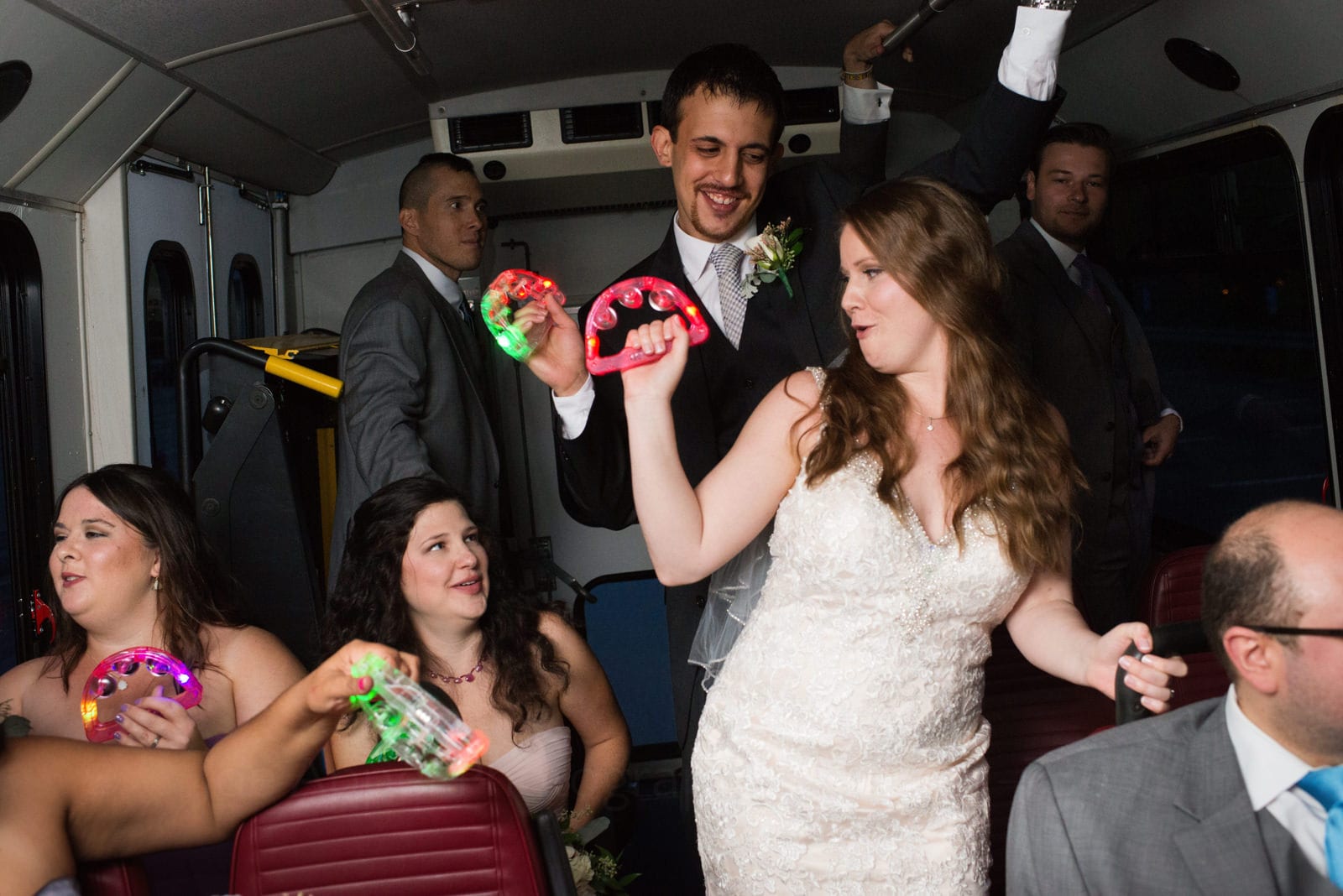 A bride and groom dance and shake lighted tambourines surrounded by their bridal party in the back of a shuttle bus during their Fall Wedding at Seven Springs.