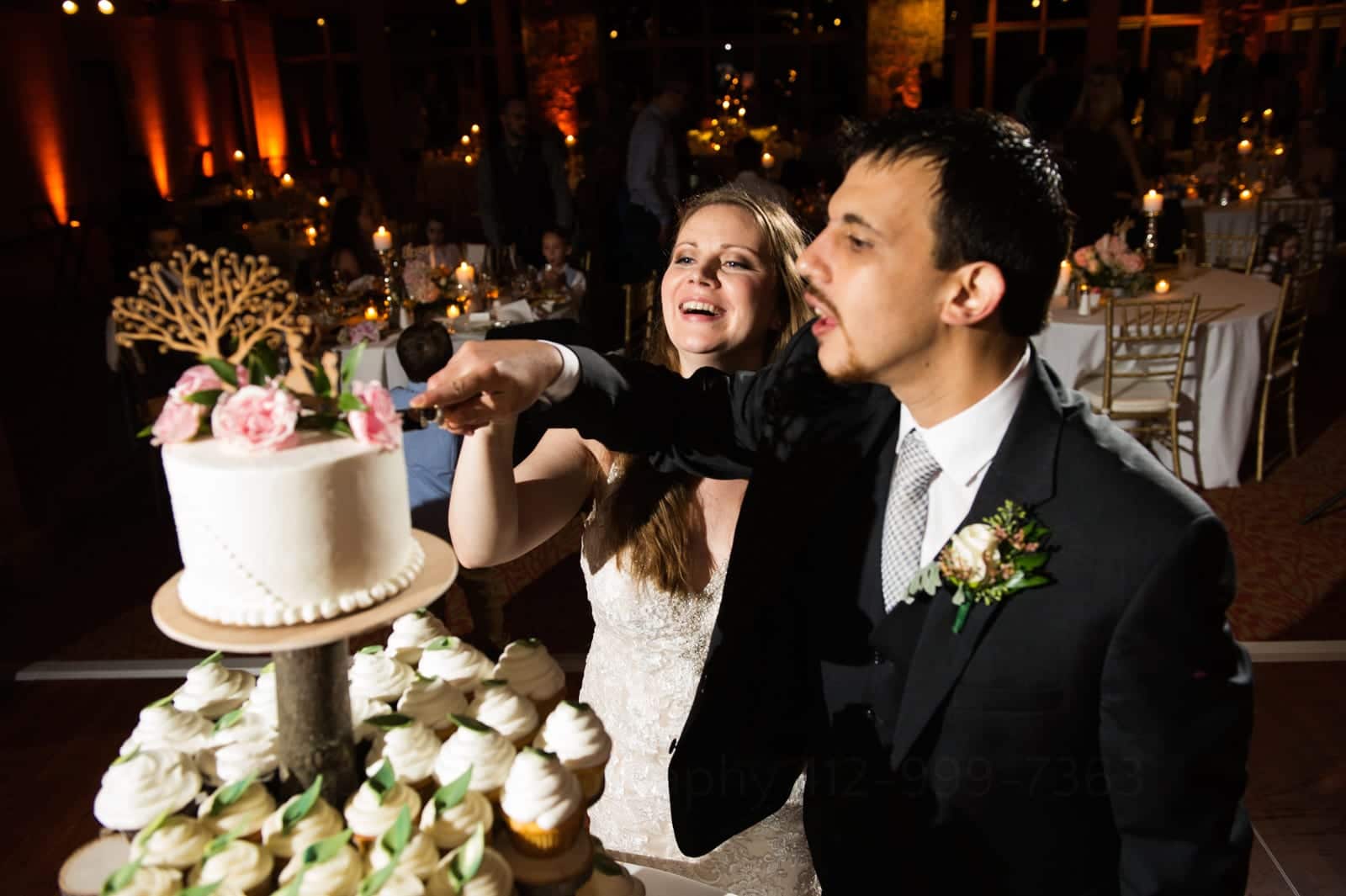 A bride and groom reach high to cut a small wedding cake atop a tower filled with cupcakes during their Fall Wedding at Seven Springs.
