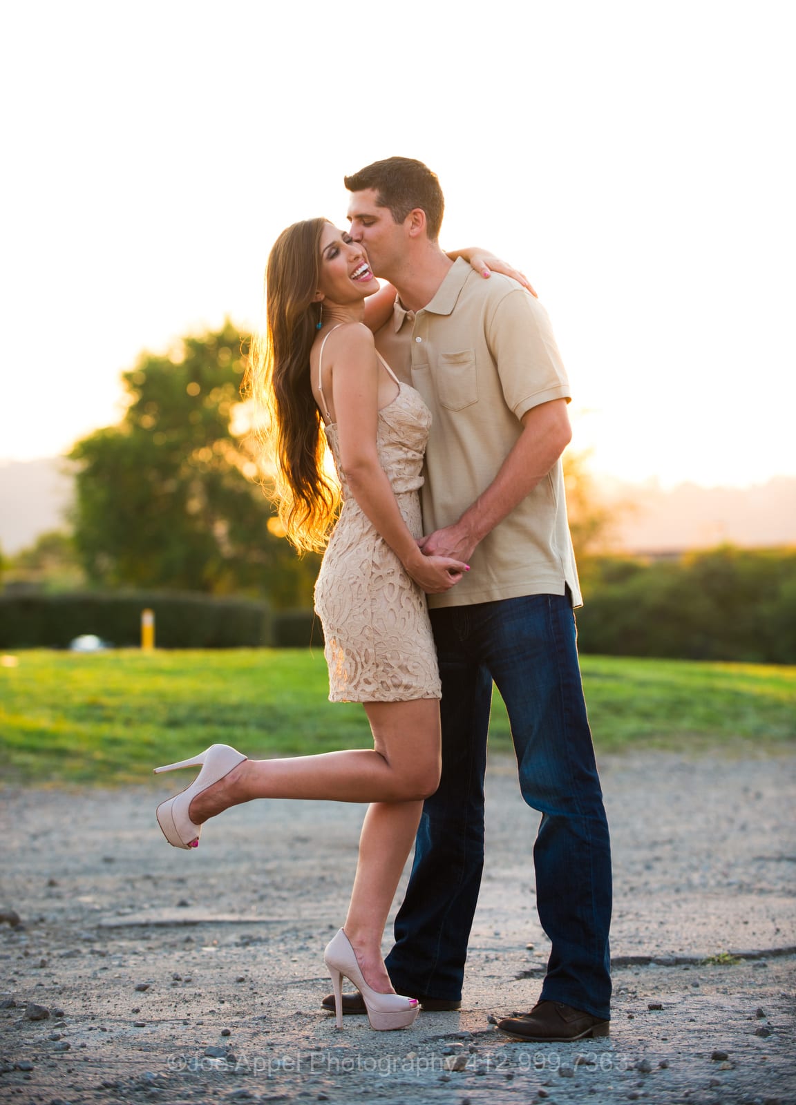 A woman in a short tan dress lifts her high heel into the air as she is held by a tall man wearing a tan shirt and blue jeans during their Pump House Engagement Photography session.