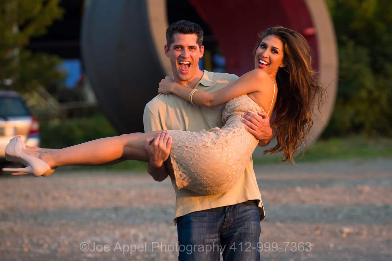 A man carries a woman wearing a tan dress as they laugh in front of an old crucible used in steel making during their Pump House Engagement Photography session.