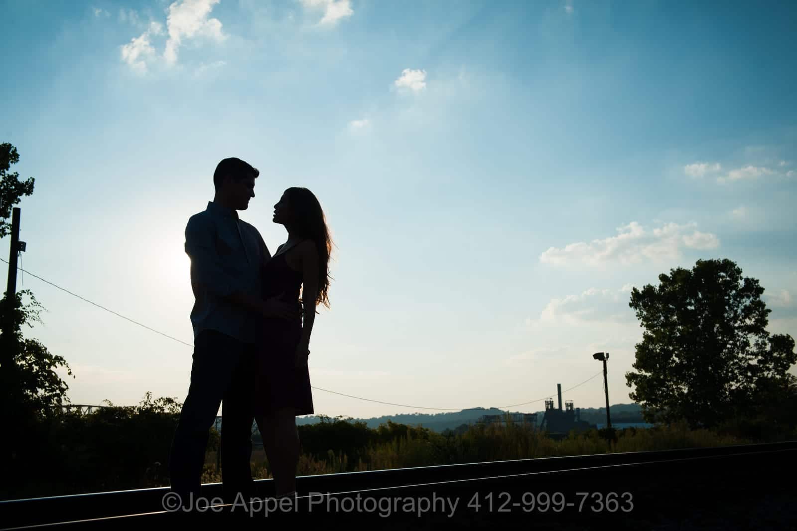 A silhouetted couple face each other as they embrace with blue skies behind them during their Pump House Engagement Photography session.