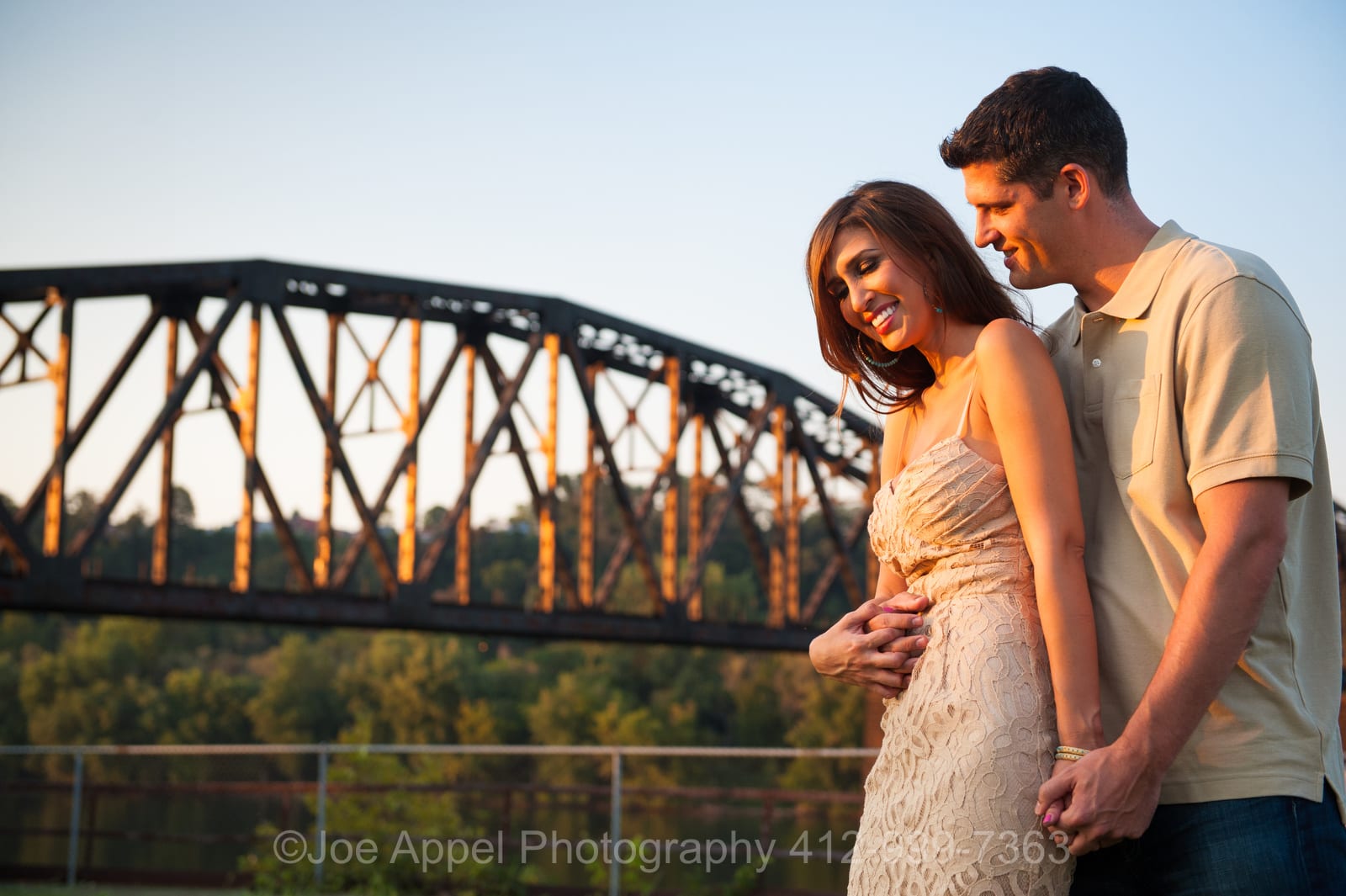 A man stands behind a woman and embraces her with an old steel bridge in the background during their Pump House Engagement Photography session.