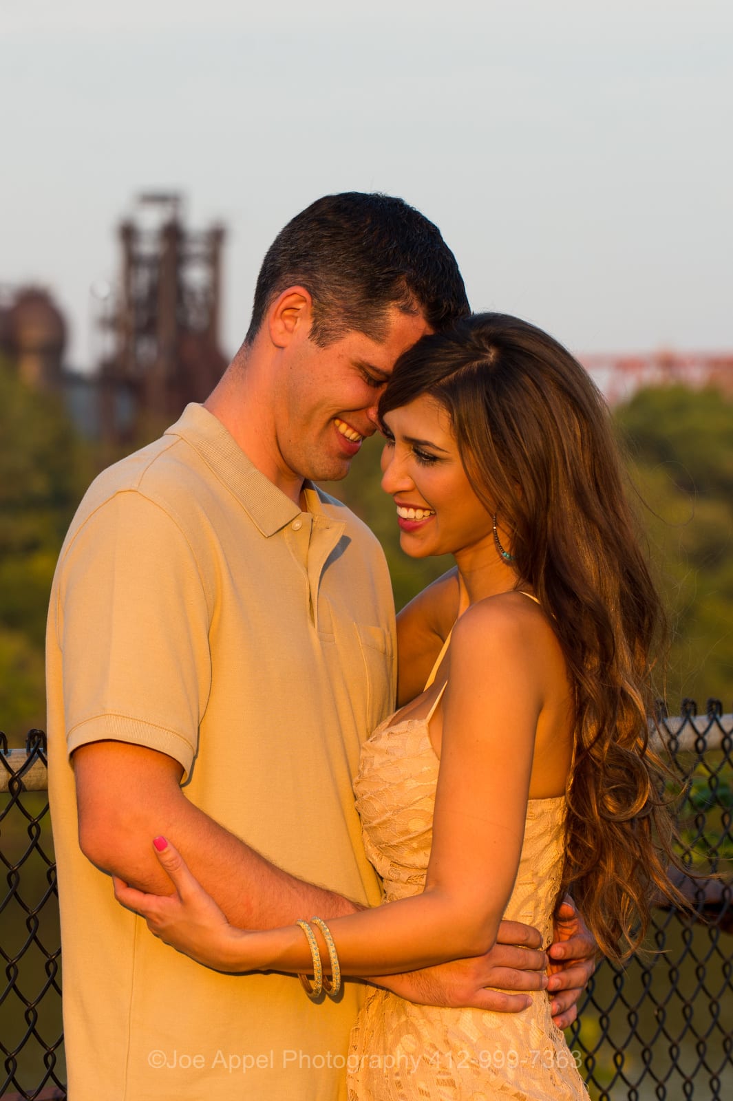 A woman and a man hold each other as the sun sets casting golden light on them in front of an old steel mill during their Pump House Engagement Photography session.