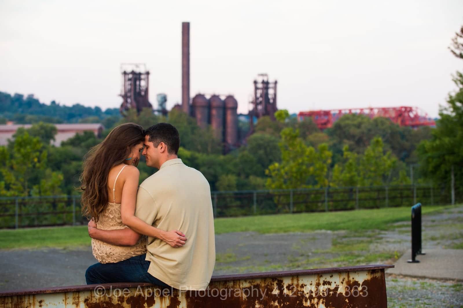 A man and a woman sit on an old i-beam with arms around each other touching foreheads with an old steel mill in the background during their Pump House Engagement Photography session.