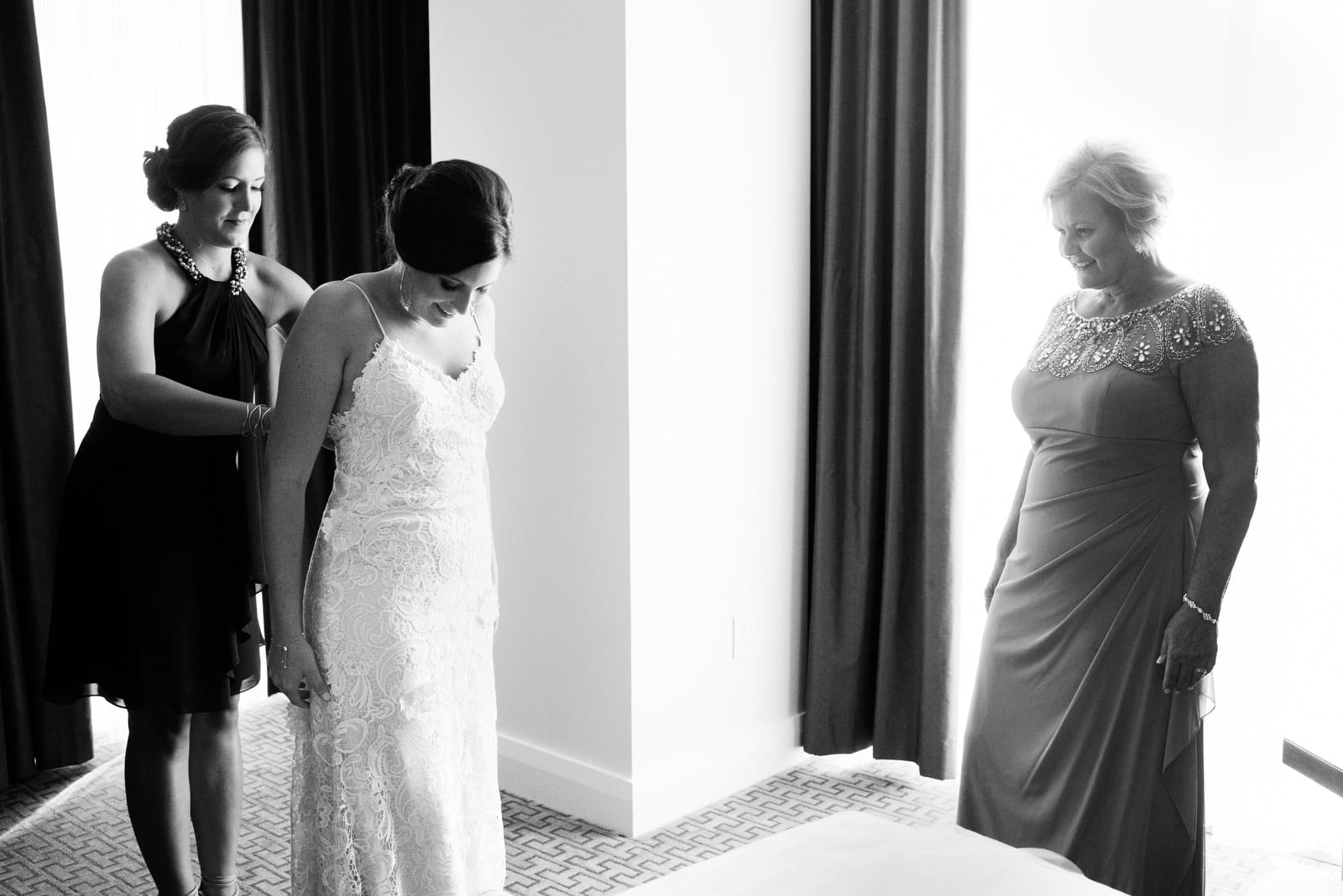 A bride looks down as her sister buttons her dress and their mother looks on before Wedding Photography at Fairmont Hotel Pittsburgh.