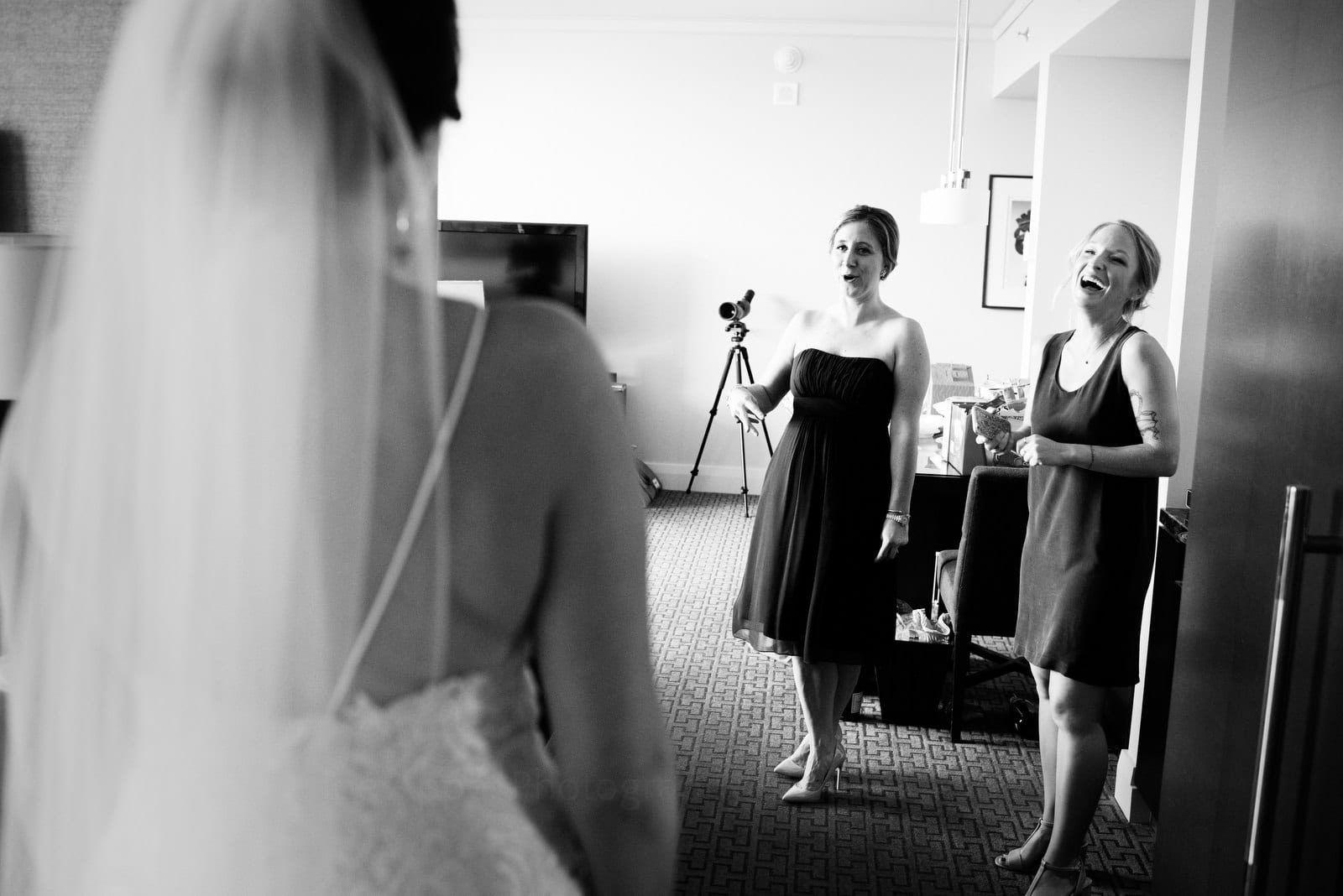 Two bridesmaids react with smiles at a bride who faces away from the camera before her Wedding Photography at Fairmont Hotel Pittsburgh.