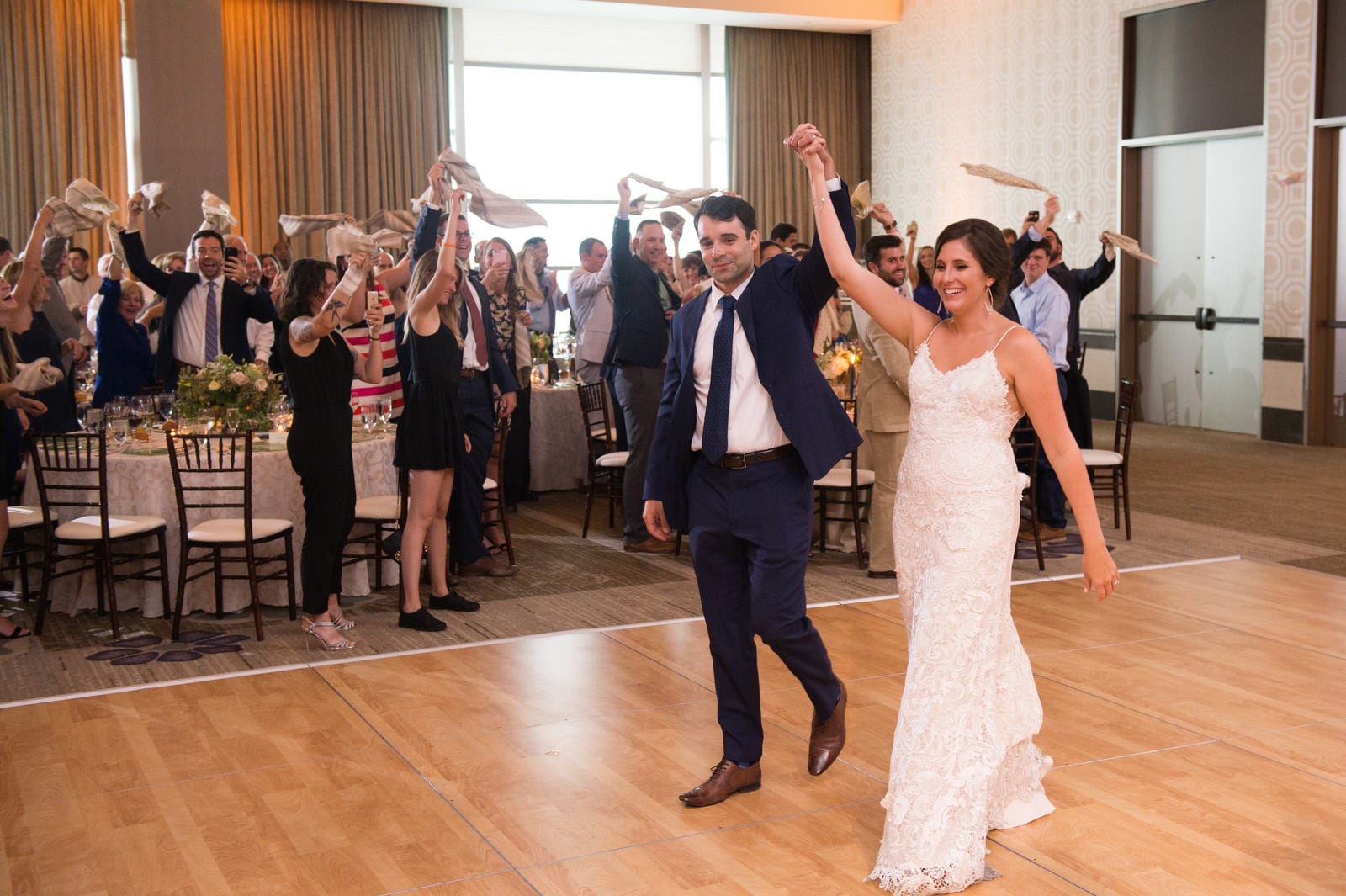 A bride and groom hold each other's hand in the air as they enter their wedding reception. Guests are waving their napkins above their heads during their Wedding Photography at Fairmont Hotel Pittsburgh.