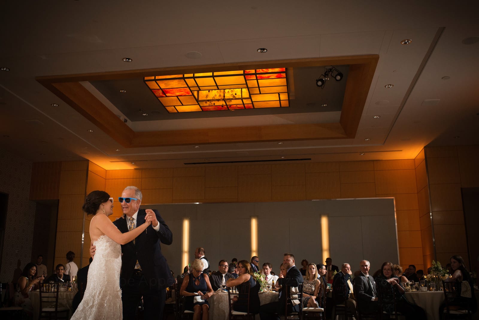 A bride and her father wear sunglasses as they dance beneath an orange and red stained glass chandelier while the guests look on Wedding Photography at Fairmont Hotel Pittsburgh.