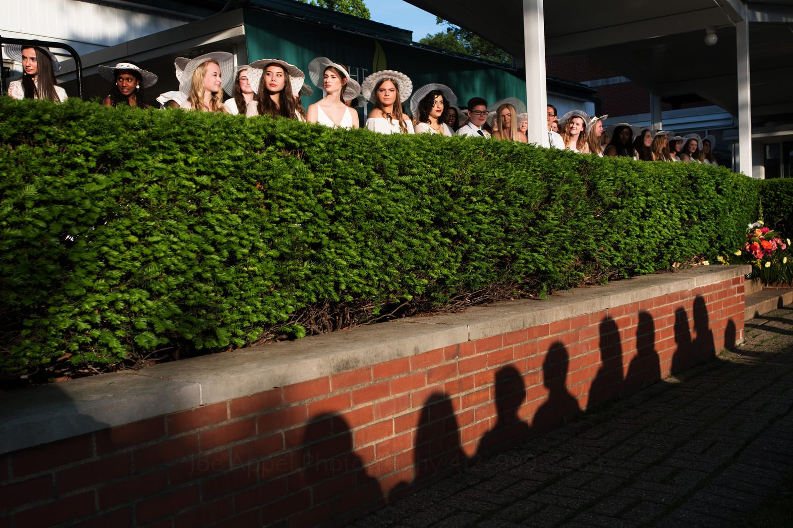 Pittsburgh editorial event photographer covers graduation ceremony at an all-girls school. Shadows of their parents appear on the brick wall beneath green hedges. The girls sit above.