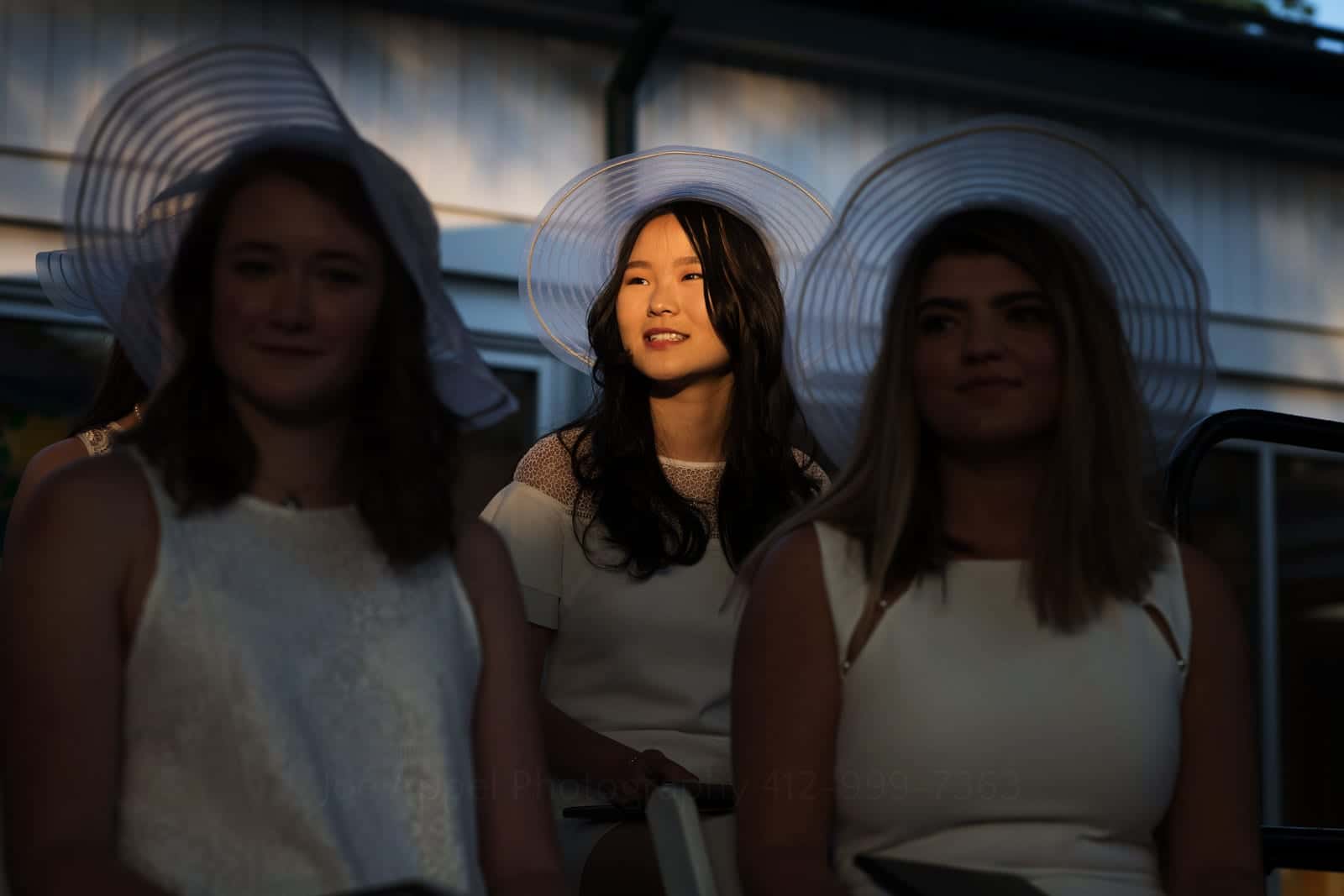 A girl wearing a white hat and dress is illuminated by a shaft of sunlight as the girls seated next to her are in shadow Pittsburgh editorial event photographer.