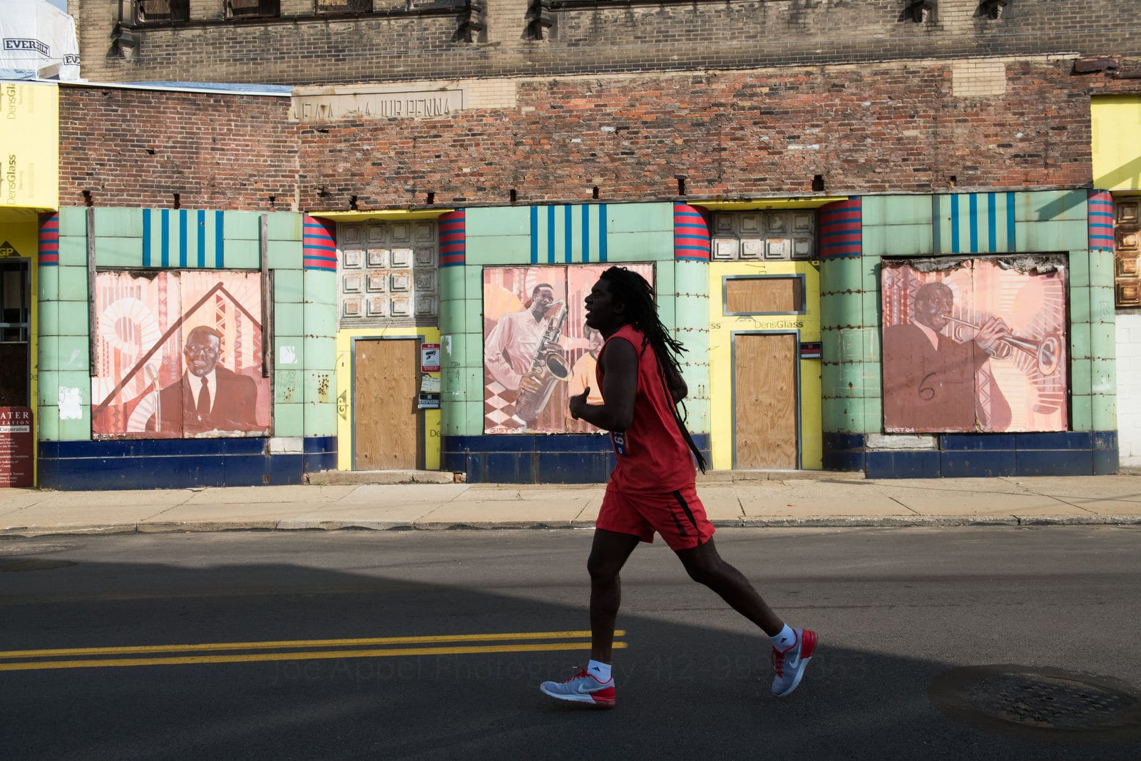 A man wearing red runs in the shadows as he passes by the New Granada Theater in the Hill District during a 5K race. Pittsburgh editorial event photographer.