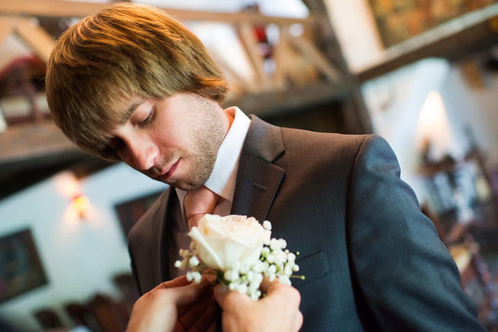 A young man with long blonde hair and a scruffy beard looks down as a pair of hands pins a white rose to his suit before his Hyeholde Wedding Pittsburgh.