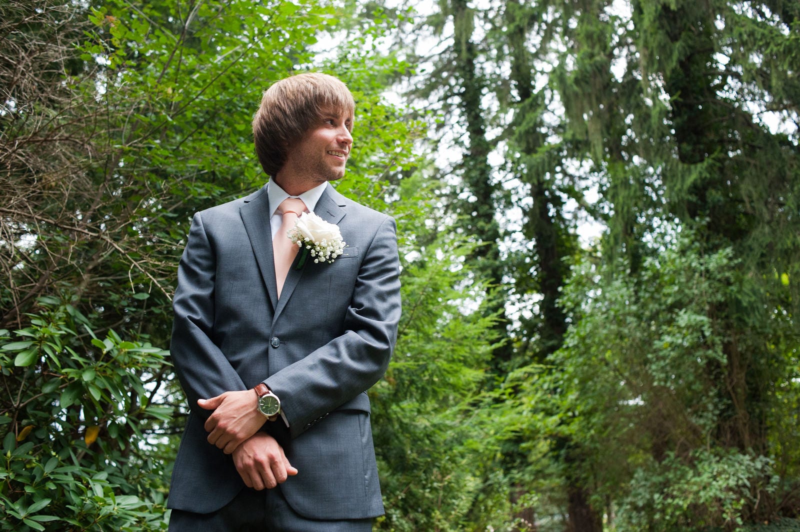 A smiling groom wearing a blue suit and pink tie stands in a clearing in the woods while watching his bride walk towards him for their Hyeholde Wedding Pittsburgh.