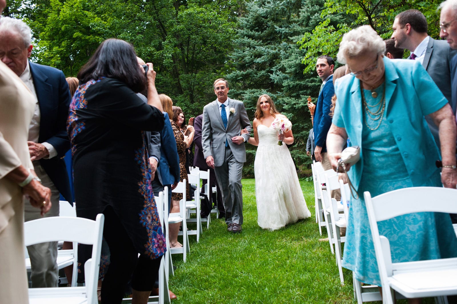 A bride walks with her father down the aisle between her guests seated on white wooden folding chairs in a wooded glen during her Hyeholde Wedding Pittsburgh.