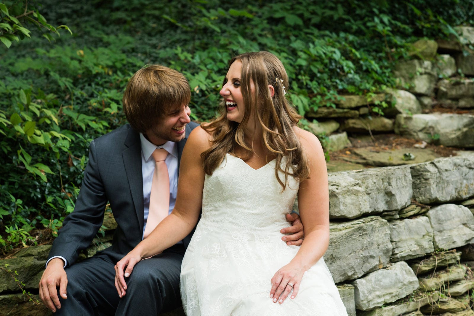 A bride and groom sit with each other on a stone wall and laugh during their Hyeholde Wedding Pittsburgh.