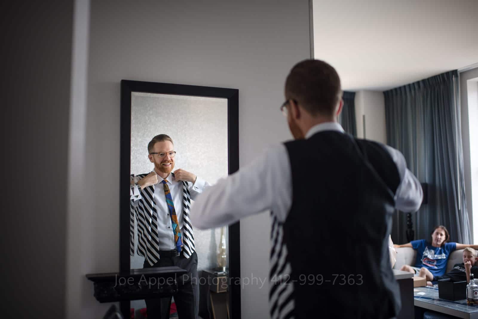 A bearded man looks in a mirror as he puts on his necktie in a hotel room. He's wearing a black and white striped vest.