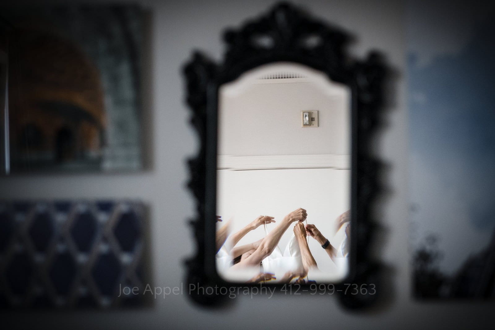 A framed mirror hanging on a wall reflects a bunch of hands holding a wedding dress as a bride gets dressed.