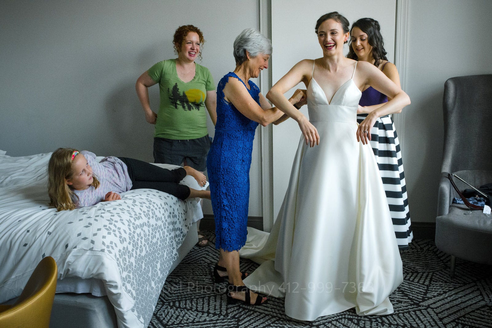 A bride smiles as her mother and a bridesmaid button up the back of the dress. A woman in a green t-shirt watches behind them and a young girl lays on a bed next to them watching.