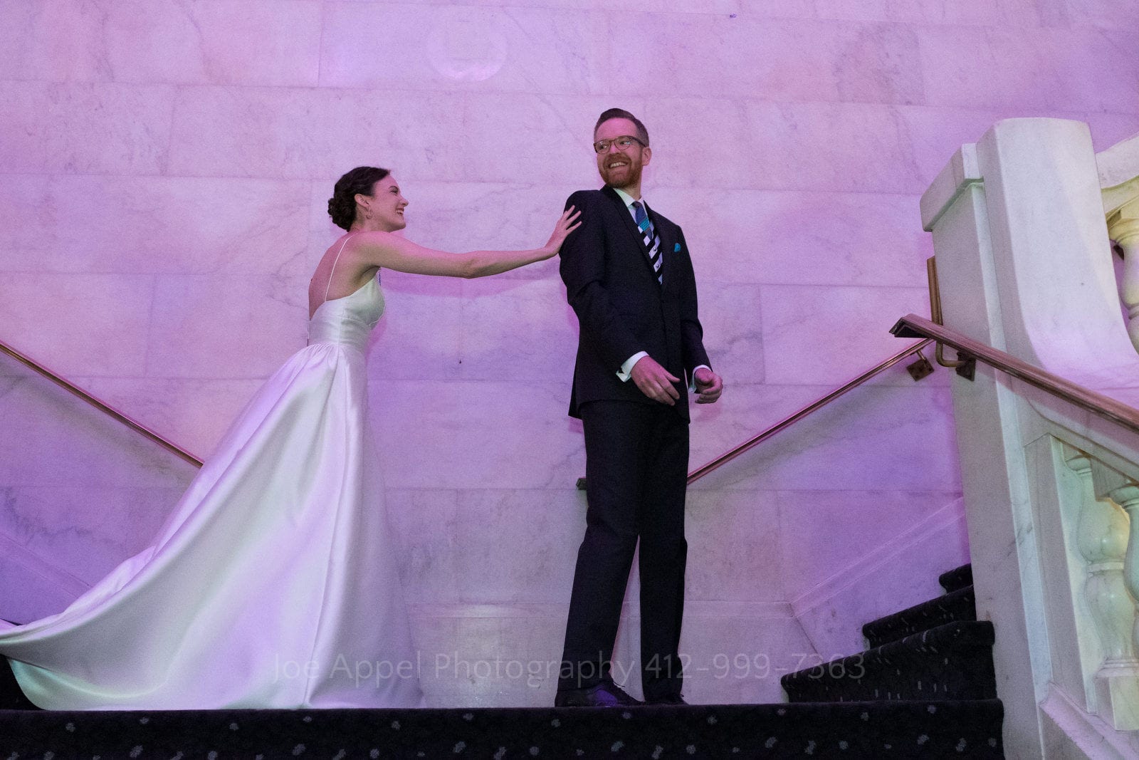 A bride reaches out to touch her groom as they stand on the landing of the grand staircase at The Renaissance hotel in Pittsburgh. The purple room light is reflected on the white marble around them.
