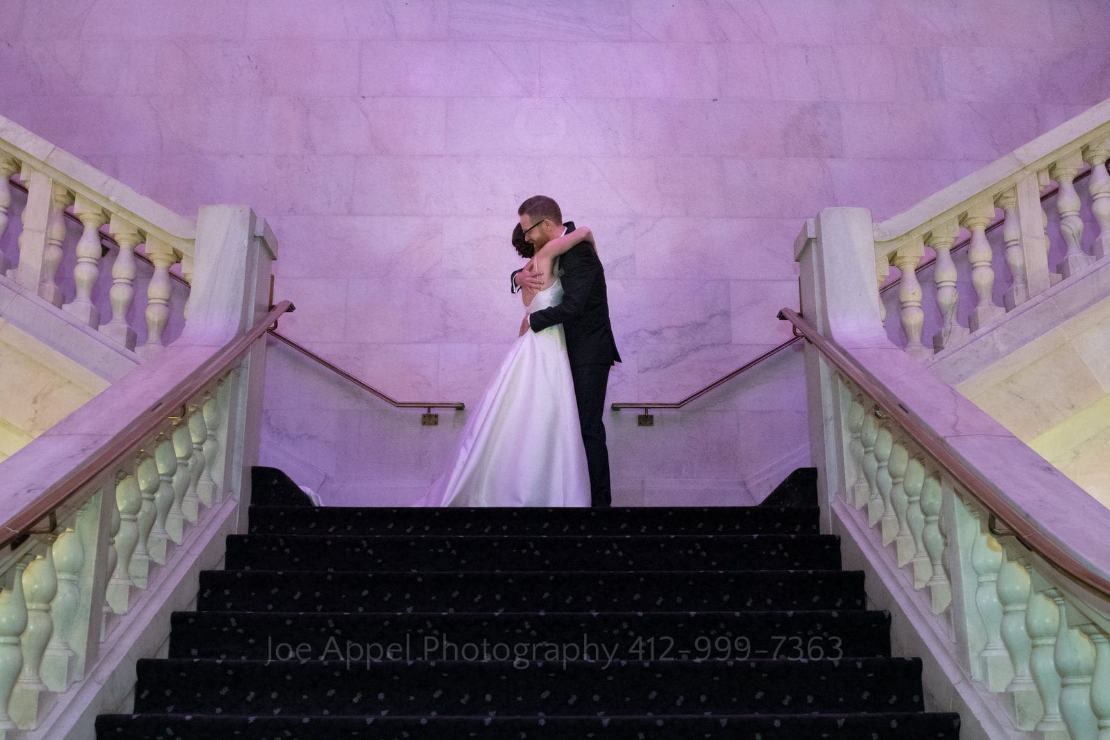 A bride and groom embrace on the landing of the grand white marble staircase at the Renaissance Hotel in Pittsburgh. Purple light reflects from the white surfaces.