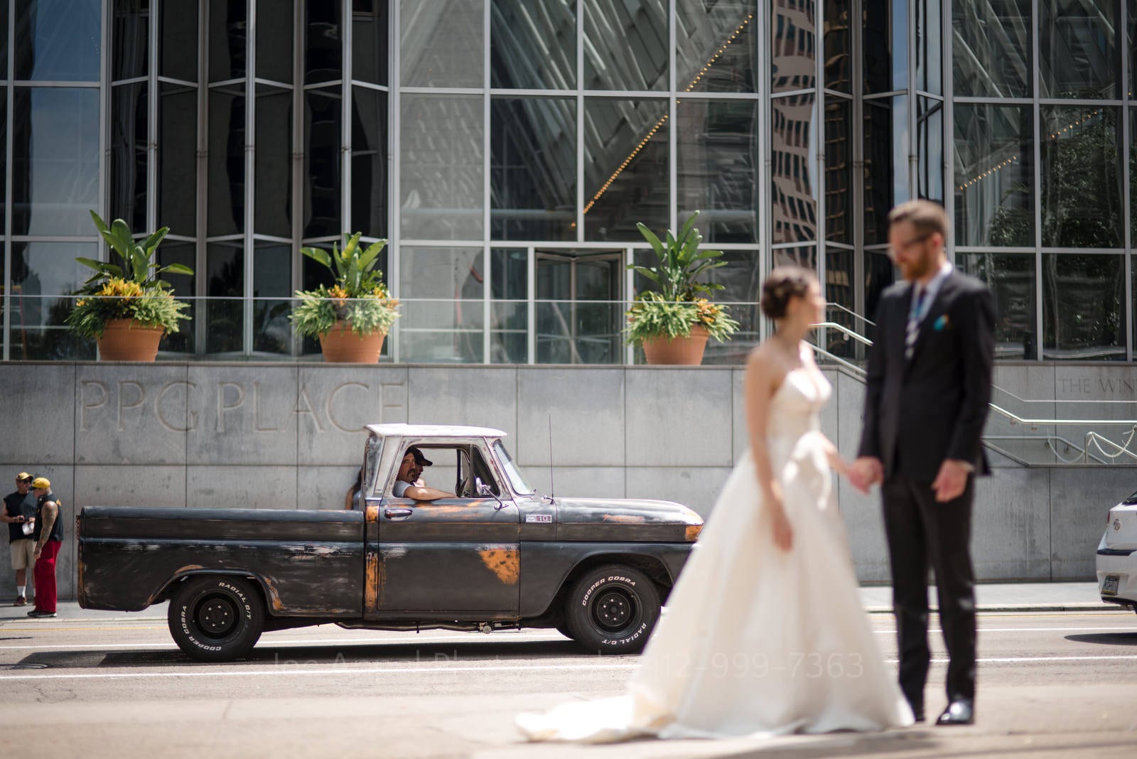 A bride and groom stand on the street as a couple of men seated in a rusty old Chevy pickup truck drive past them on Stanwix Street in downtown Pittsburgh.