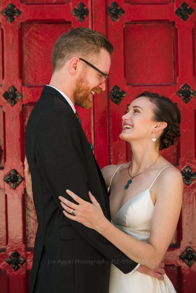 A bride and groom embrace and smile at each other in front of the red wooden door on the Stephen Foster Memorial in Pittsburgh.