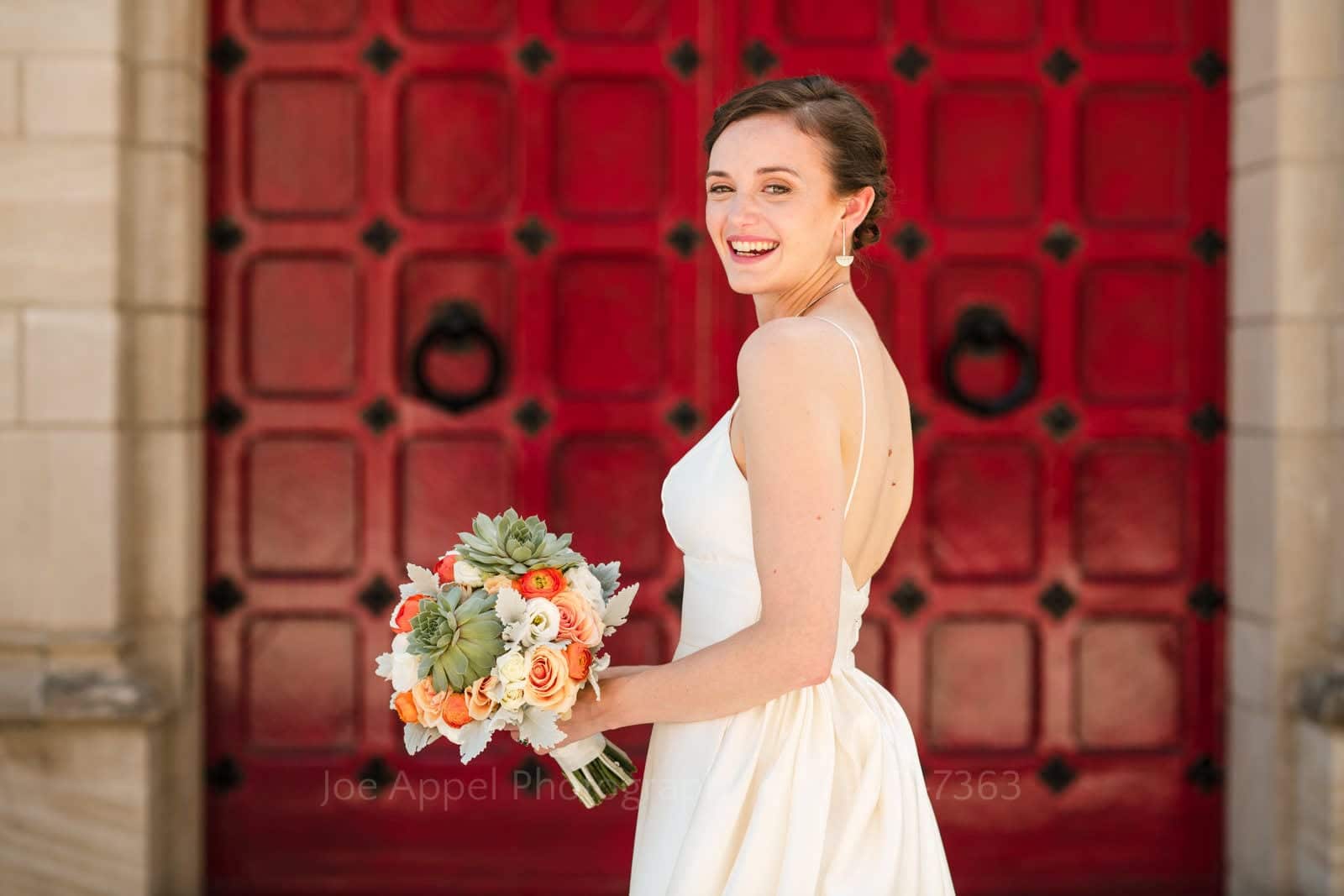 A smiling bride holds her bouquet as she looks over her shoulder while standing in front of a set of red wooden doors at the Stephen Foster Memorial.