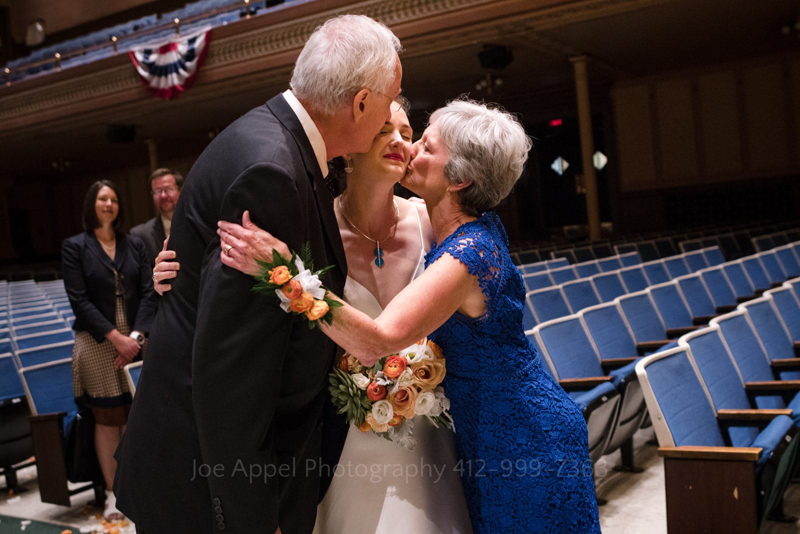 A bride stands in an auditorium and is kissed on each of her cheeks by her parents during Soldiers and Sailors Wedding Photos.
