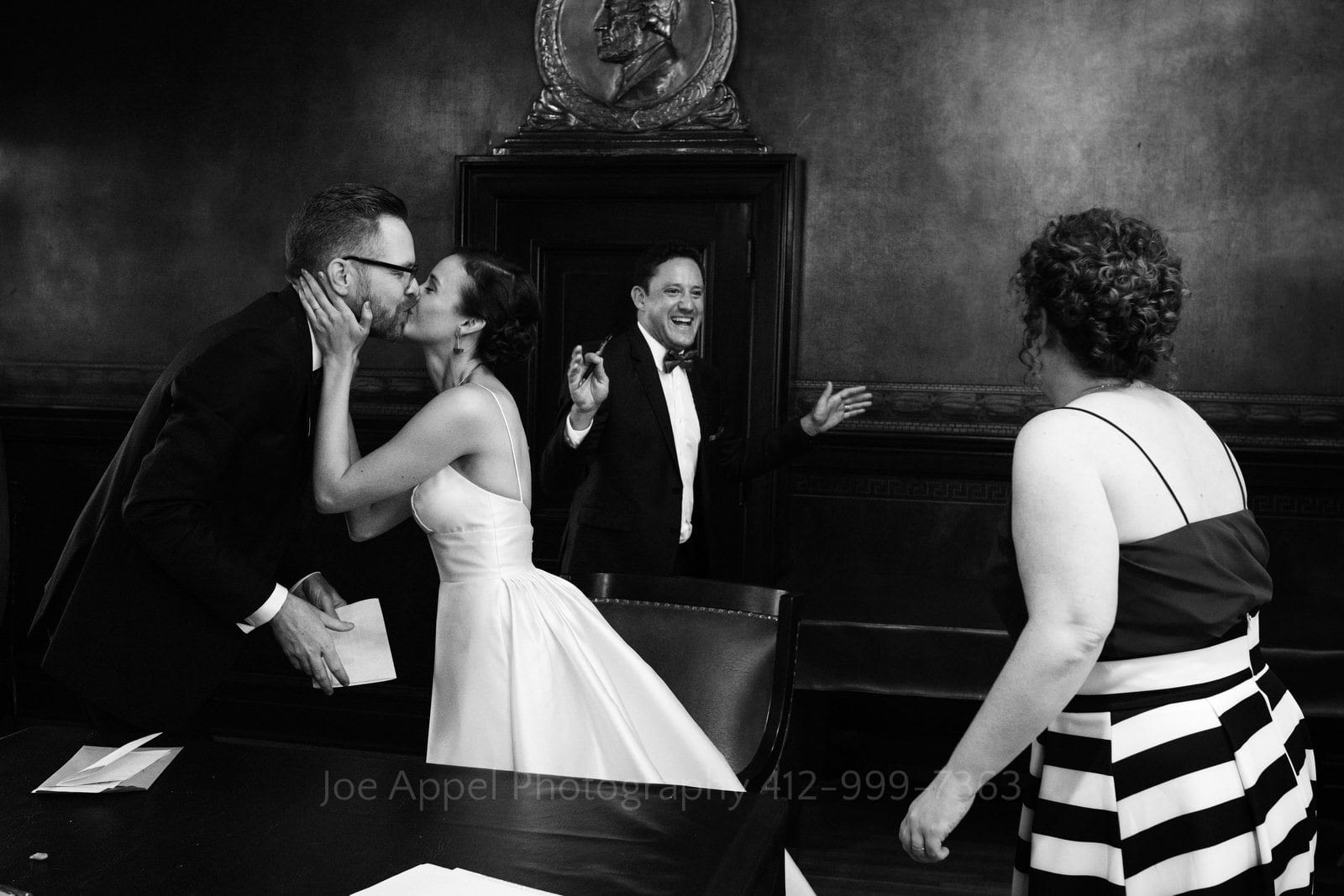 A bride and groom kiss after signing their wedding license while their witnesses run towards each other smiling Soldiers and Sailors Wedding Photos.