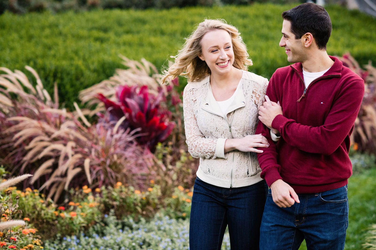 A couple walk past some brightly colored plants arm in arm outside of Phipps Conservatory and Botanical Garden. She's wearing a white jacket and he's wearing a dark red sweater.