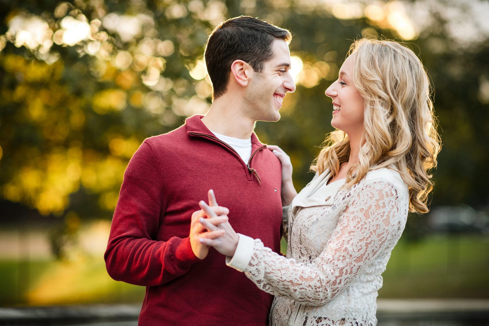 A dark haired young man wearing a red sweater holds a blonde woman wearing a white jacket with the sunset shining through the trees behind them during their University of Pittsburgh engagement portraits.