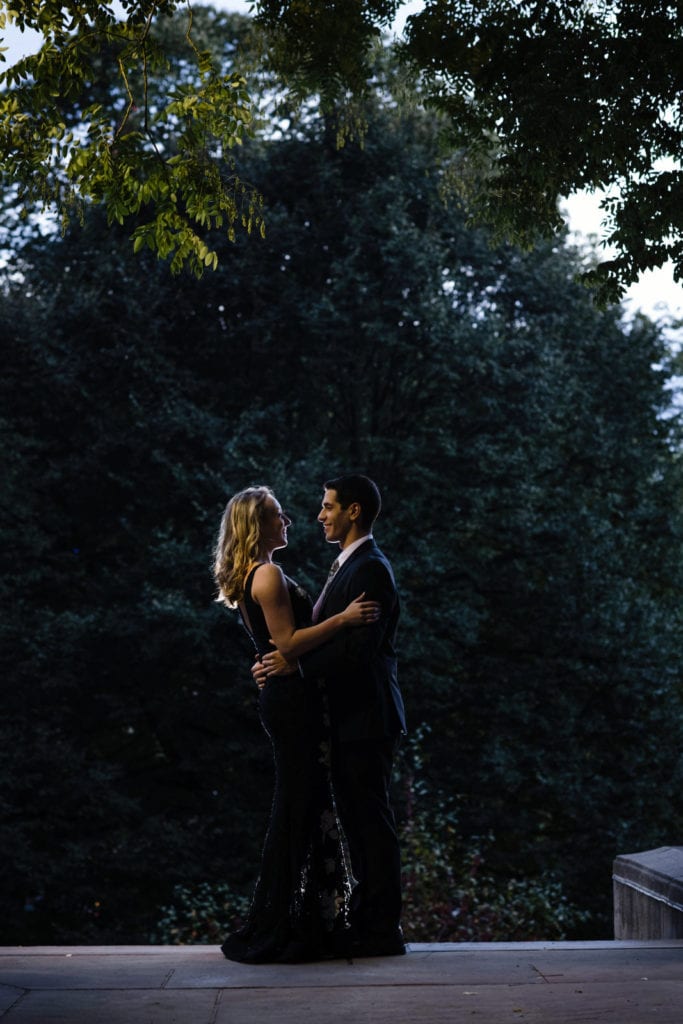 A young couple dressed in evening wear embrace at the end of a path with a tree in the background near the Cathedral of Learning during their University of Pittsburgh engagement portraits session.