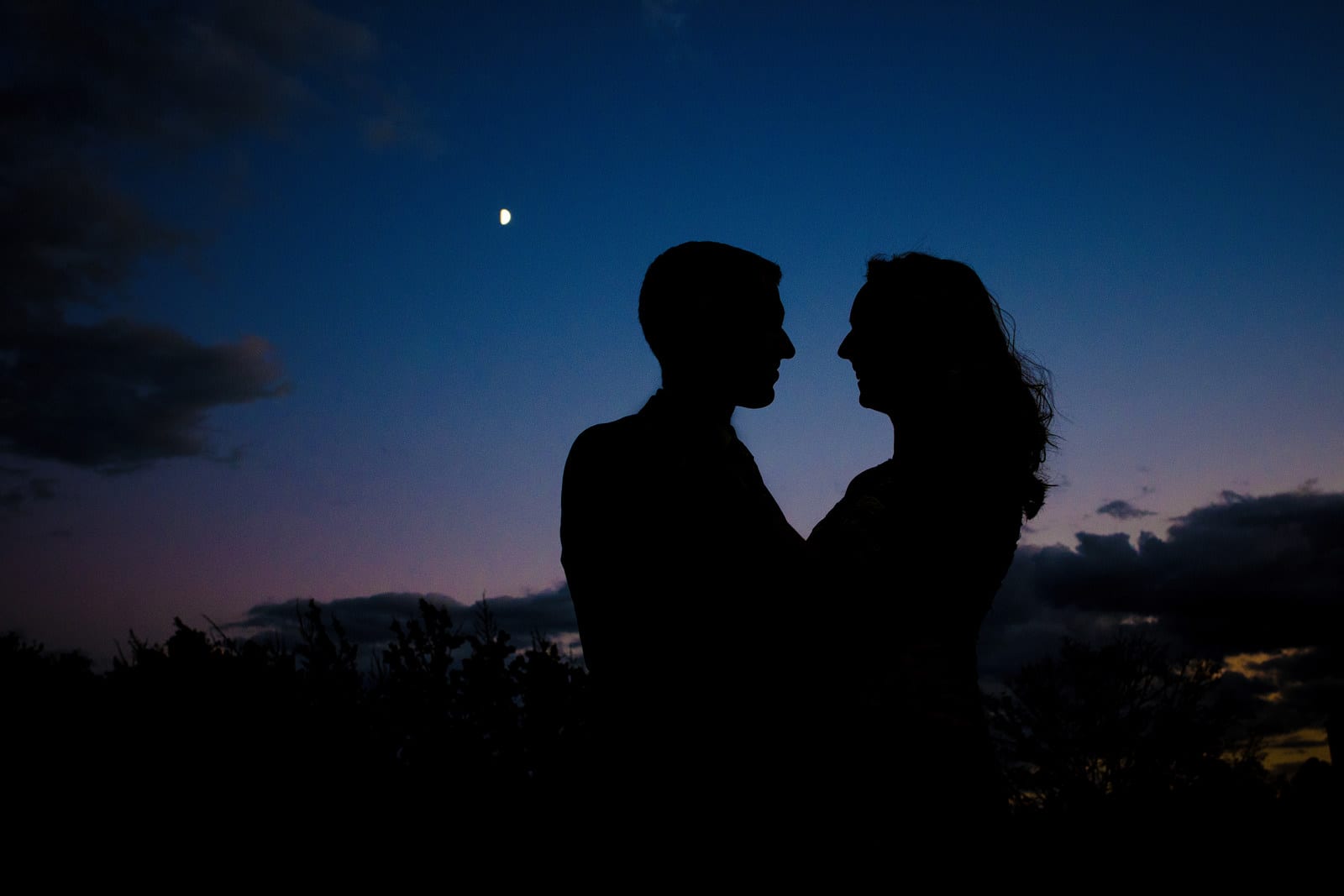 Silhouette of a couple with deep blue sky behind them and a speck of light that is the moon on the campus of the University of Pittsburgh engagement portraits.
