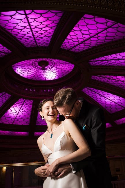 A bride laughs as her groom kisses her on the shoulder beneath the purple skylight in the lobby of the Renaissance Hotel Pittsburgh.