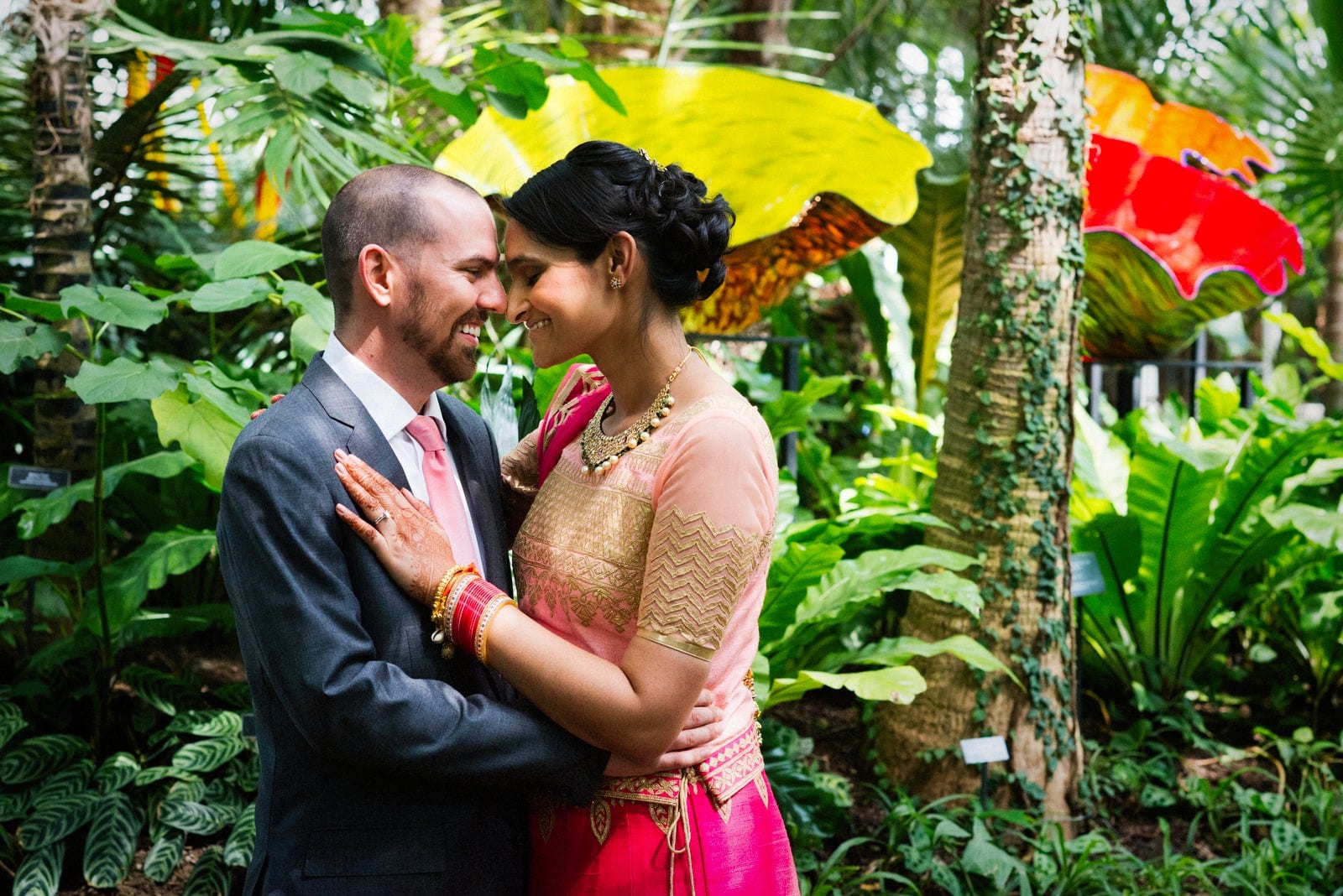 a bride dressed in a sari and groom with a grey suit and pink tie embrace in front of plants and Chihuly glass at their renaissance phipps south asian wedding