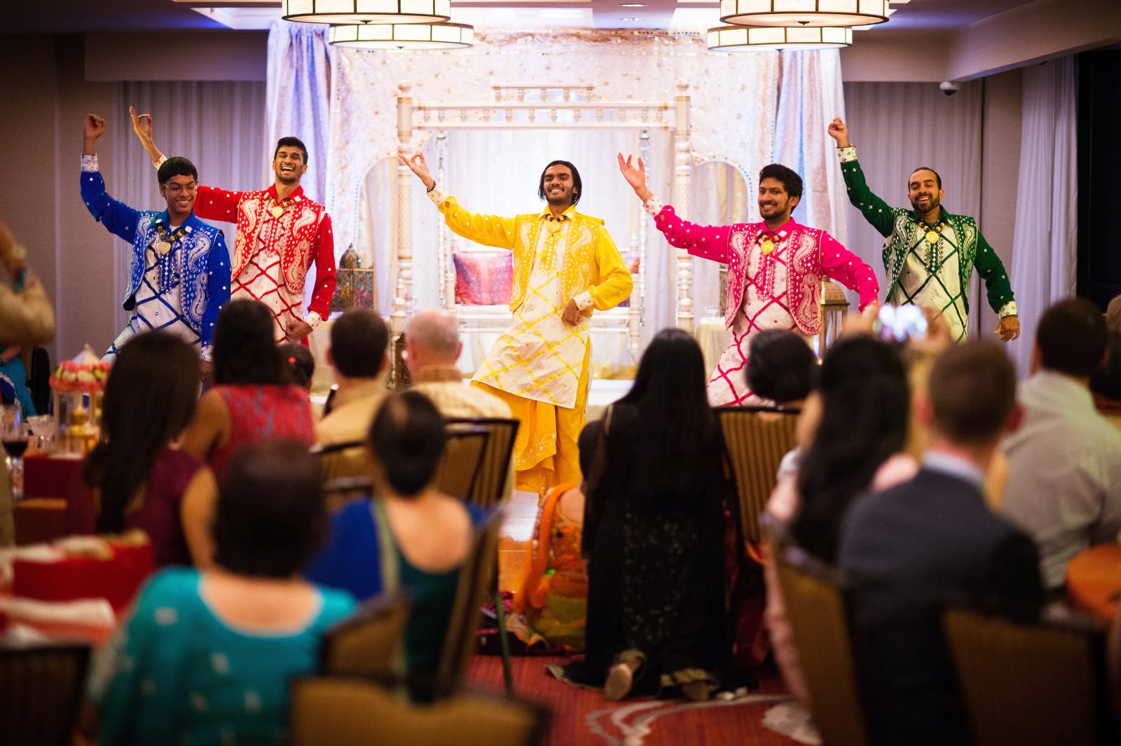 Four men wearing brightly colored clothes dance in front of guests during a Renaissance Phipps South Asian Wedding.