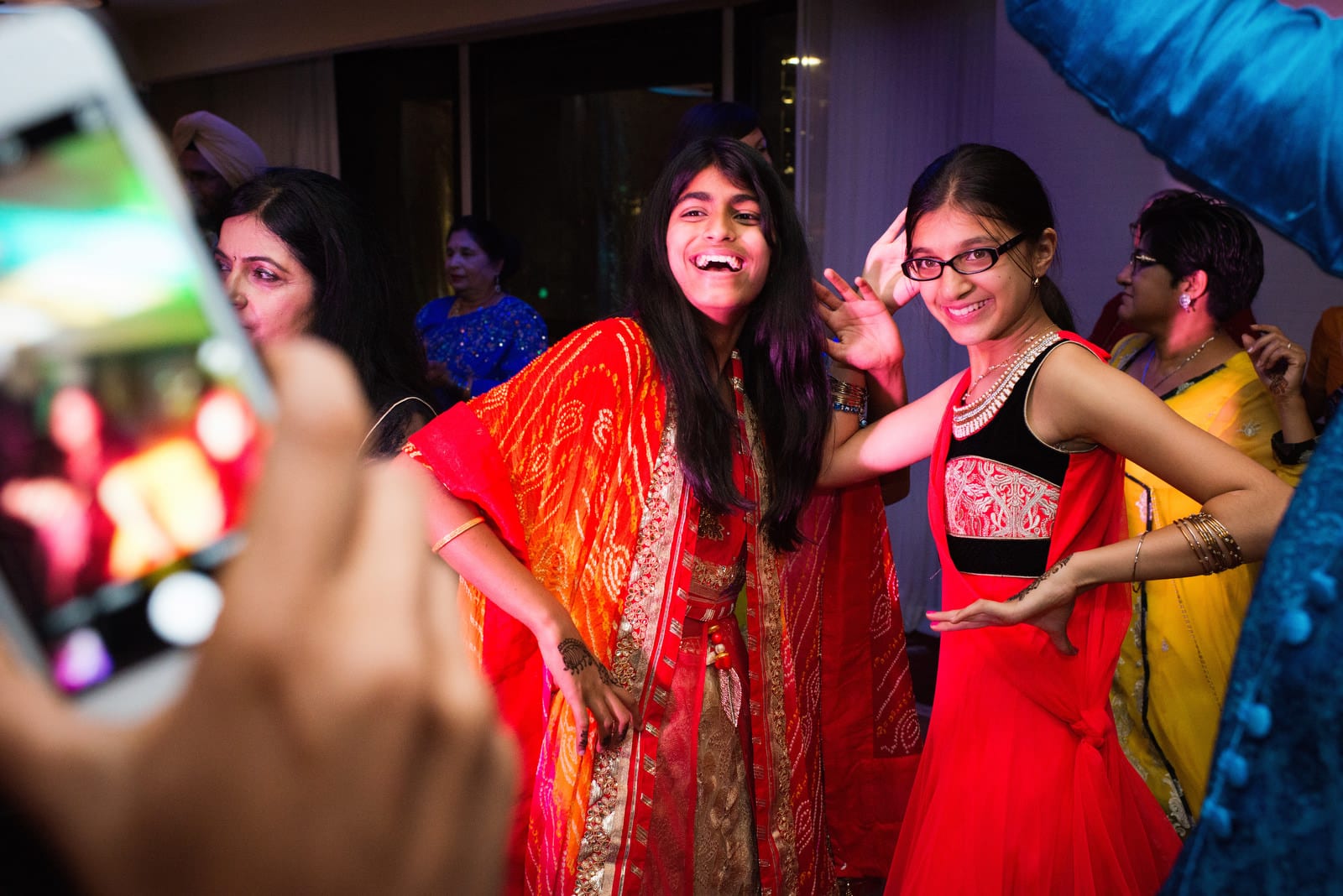 Two girls wearing bright colored clothing dance together as they are seen through the lens of a cell phone camera during a Renaissance Phipps South Asian Wedding.