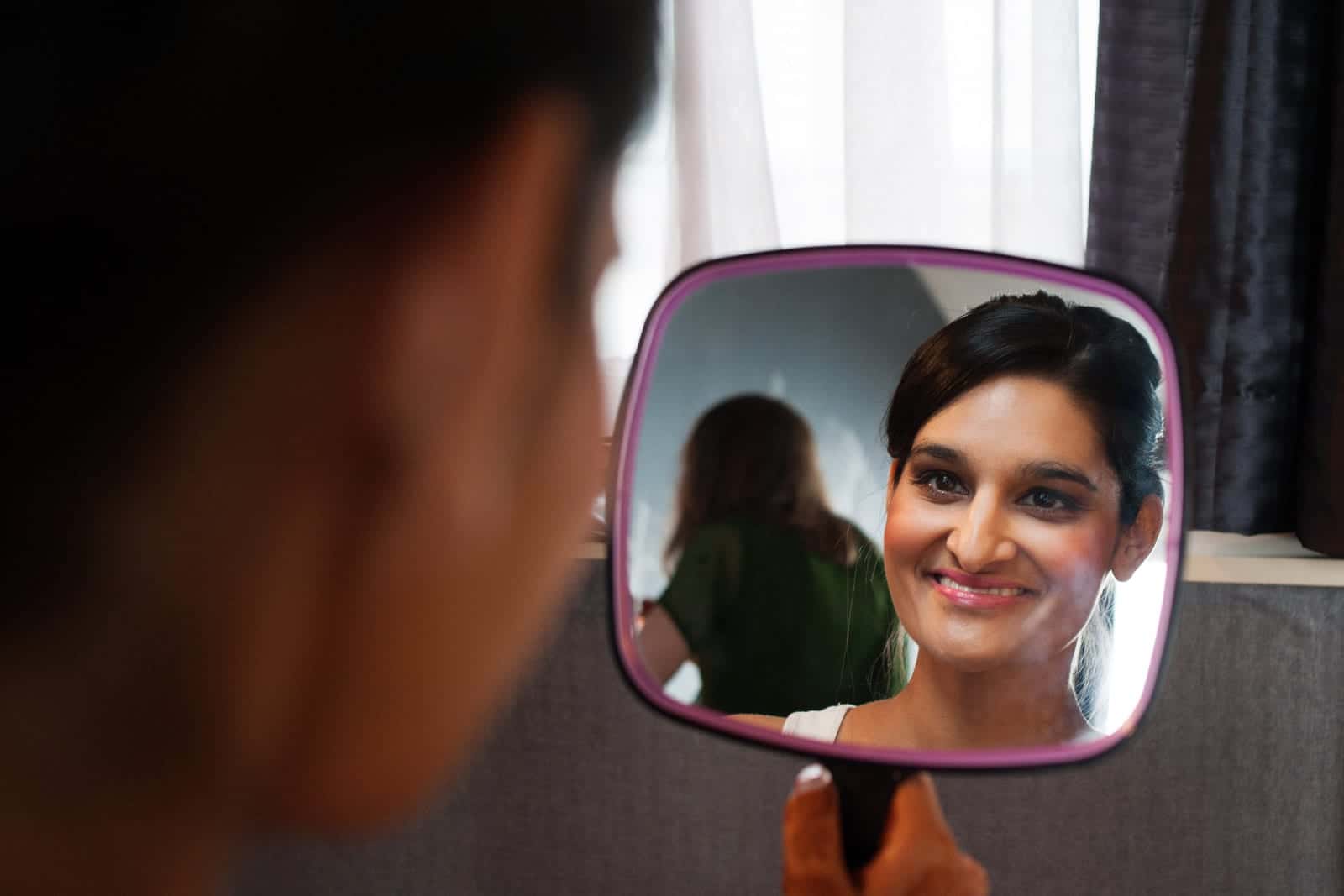 A bride smiles as she looks at her makeup in a hand mirror before her Renaissance Phipps South Asian Wedding.
